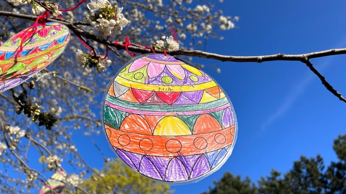 A colorful handmade Easter egg decoration hanging from a blooming branch against a clear blue sky.