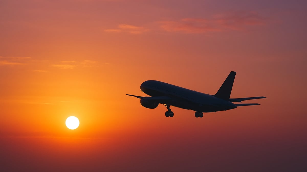 A passenger airplane takes off, silhouetted against a bright orange sunrise sky with a glowing sun.