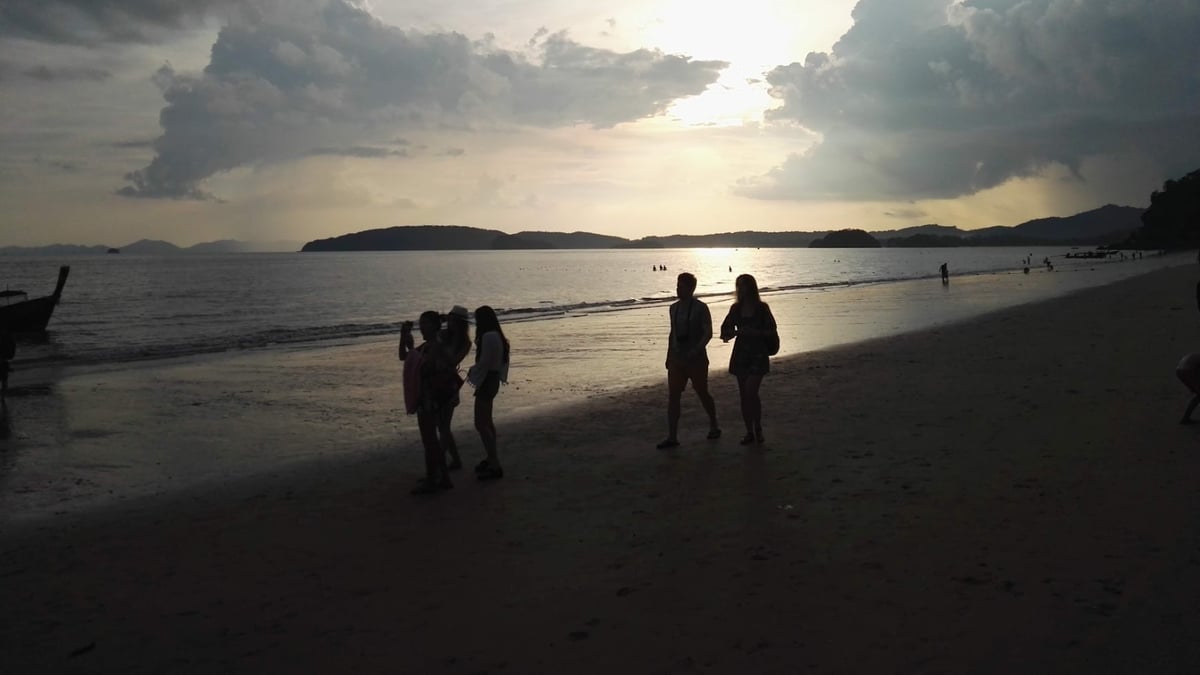 Silhouetted people walk along a sandy beach with dramatic sunset clouds.