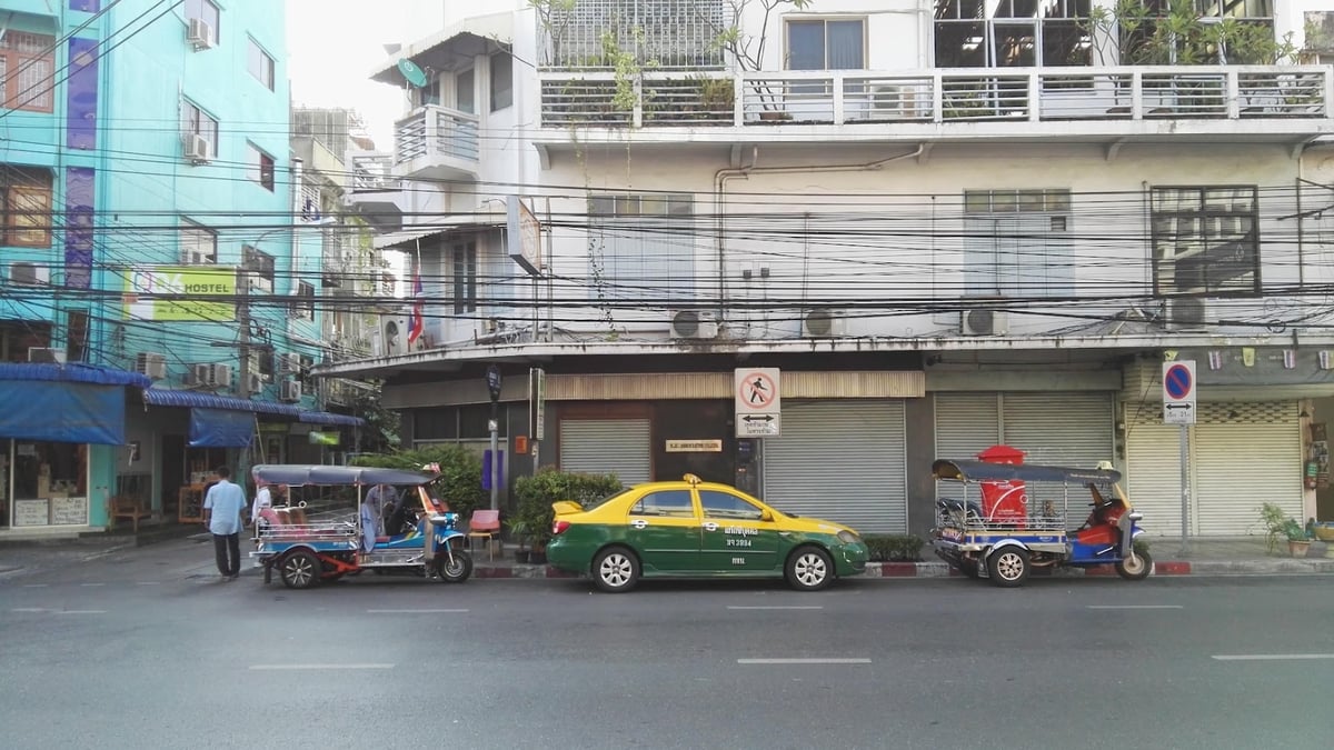 Tuk tuks and a green-yellow taxi parked along a city street with buildings in the background.