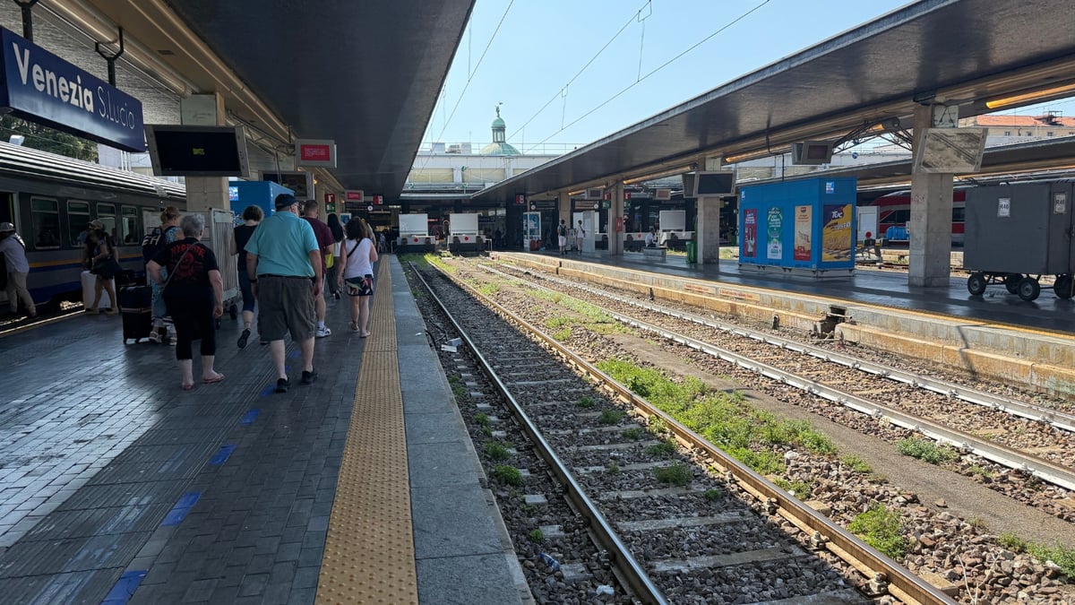 Passengers walk beside train tracks under covered platforms at Venezia S. Lucia station on a sunny day.