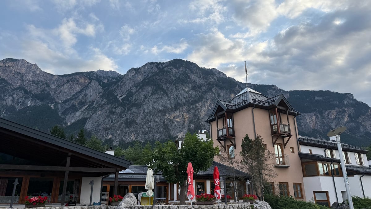 A hotel with balconies and umbrellas sits at the base of steep rocky mountains under a cloudy sky.