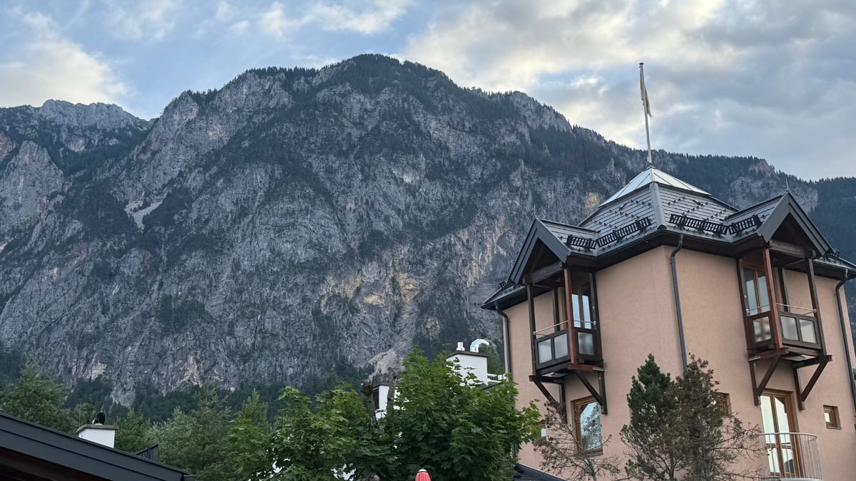 A hotel tower with wooden balconies stands before steep rocky mountains under a partly cloudy sky.