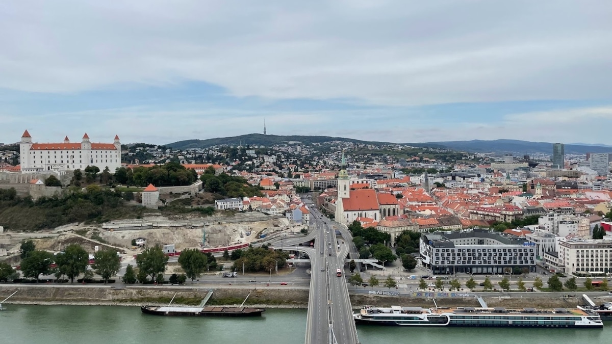 Aerial panoramic view of a historic city featuring a castle, a bridge over a river, and a mix of modern and traditional architecture.
