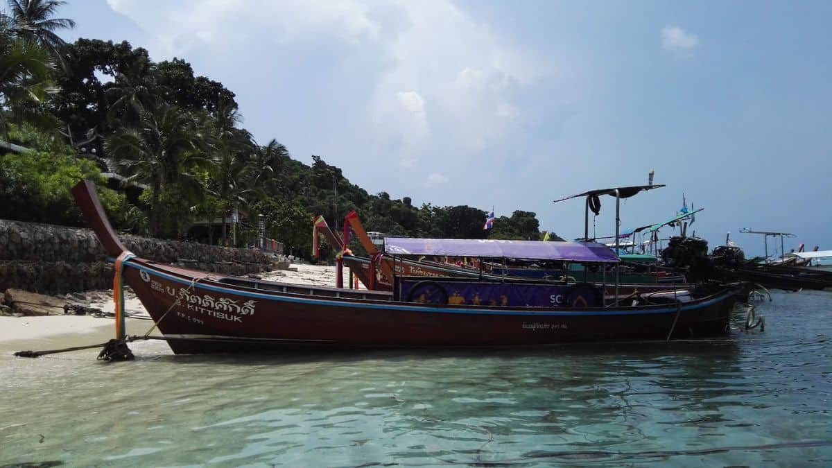 A traditional Thai long-tail boat anchored on a sandy beach with clear turquoise water and lush greenery in the background.