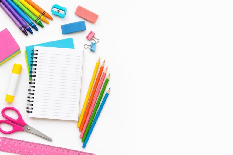 colorful crayons, pencils, erasers, scissors, and notebooks arranged on white desk with blank notebook page