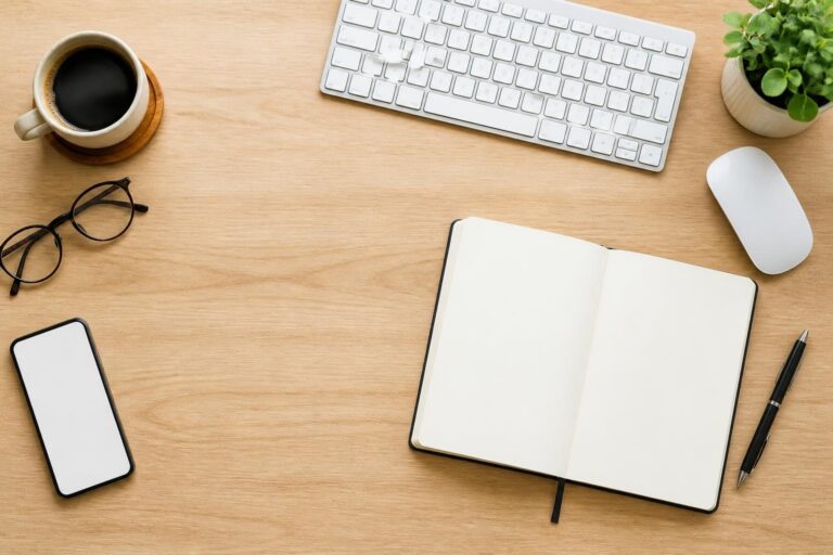Top view of a wooden desk with a keyboard, mouse, open blank notebook, pen, glasses, smartphone, coffee cup, and small plant