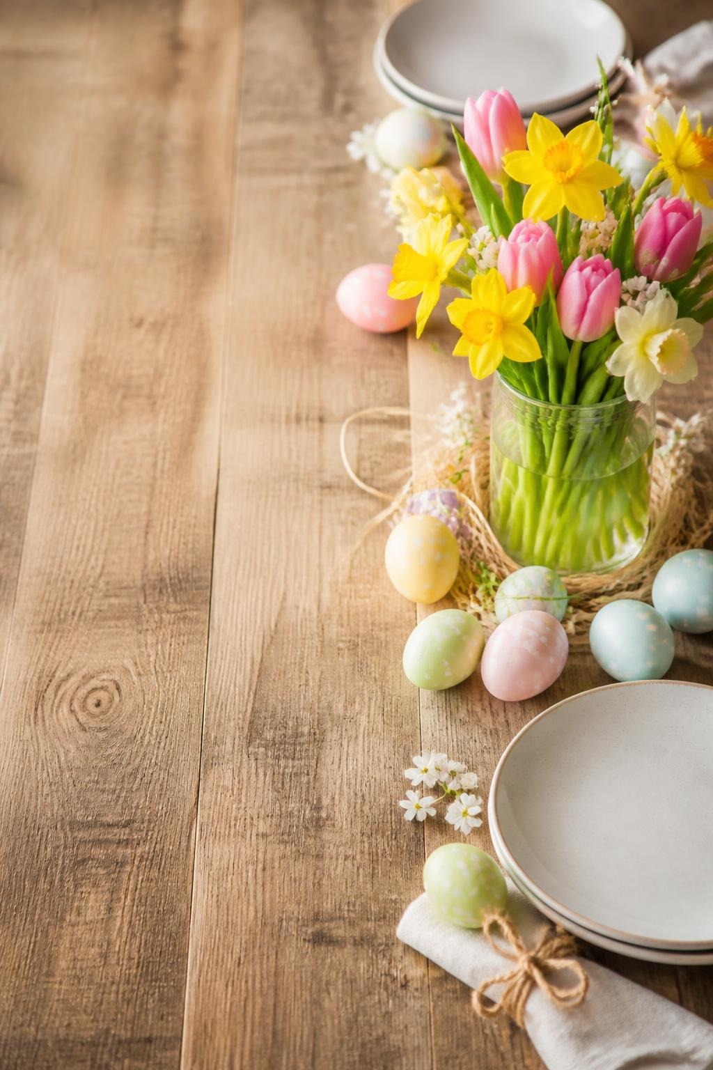 Rustic Easter table setting with pink tulips, yellow daffodils, pastel Easter eggs, and stacked plates on wooden surface.
